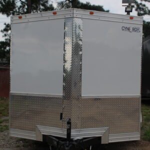 A white enclosed trailer with two axles and four wheels parked on a gravel lot with patches of grass. The trailer is positioned in front of a line of tall pine trees under a cloudy sky.