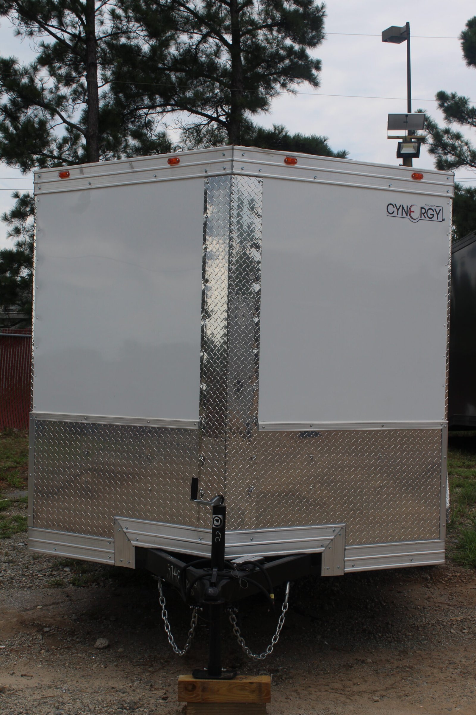 A white enclosed trailer with two axles and four wheels parked on a gravel lot with patches of grass. The trailer is positioned in front of a line of tall pine trees under a cloudy sky.