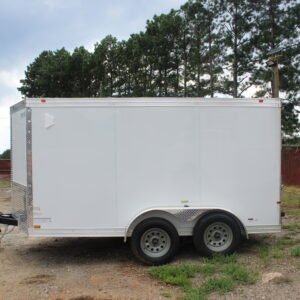 A white enclosed trailer with two axles and four wheels parked on a gravel lot with patches of grass. The trailer is positioned in front of a line of tall pine trees under a cloudy sky.