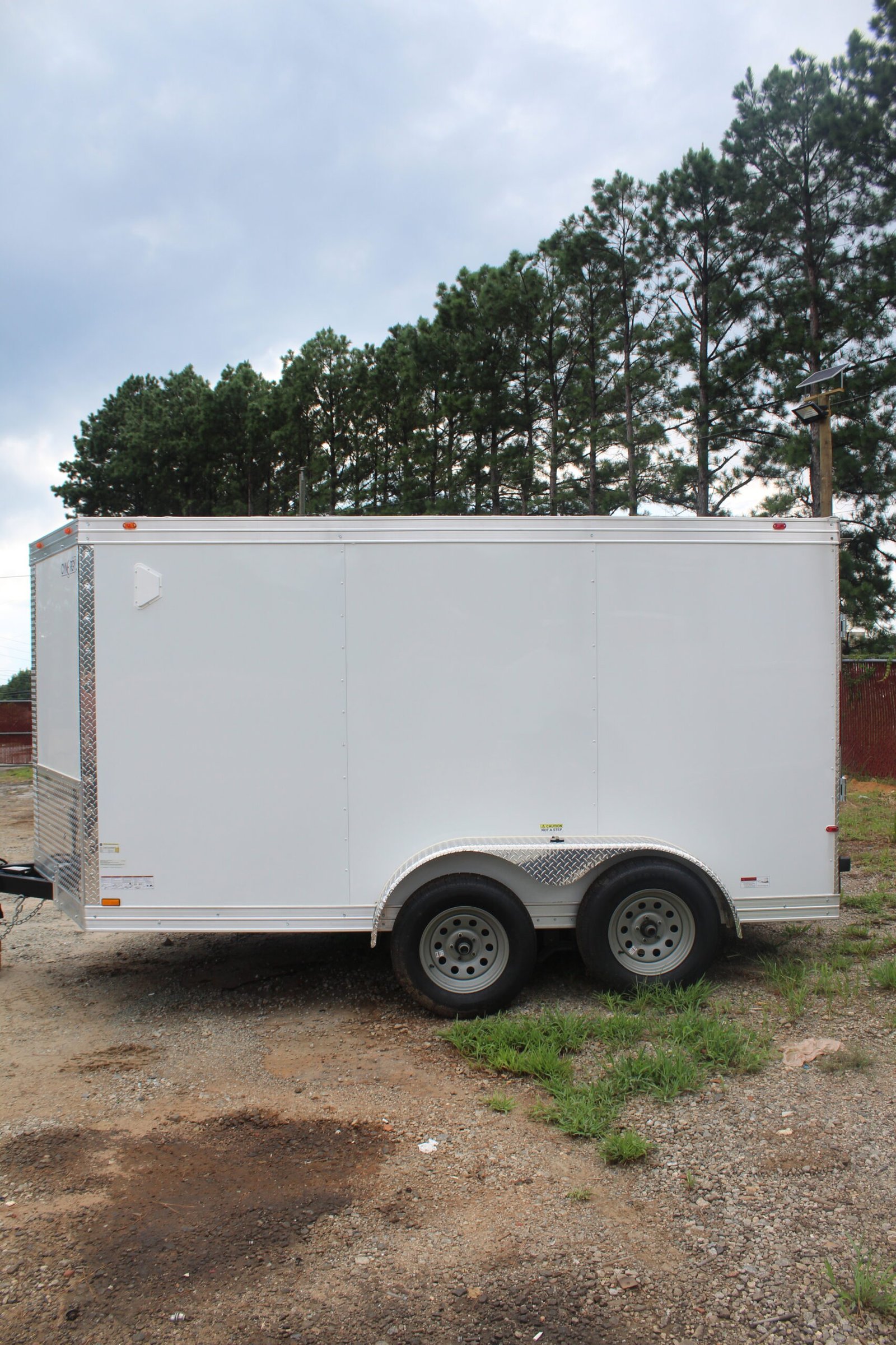 A white enclosed trailer with two axles and four wheels parked on a gravel lot with patches of grass. The trailer is positioned in front of a line of tall pine trees under a cloudy sky.