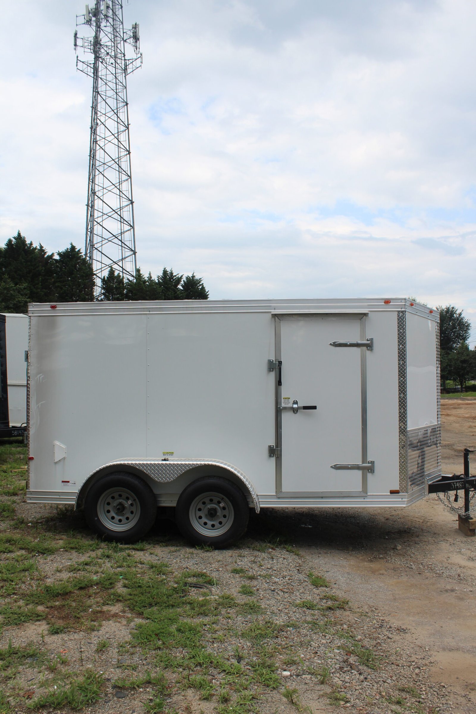 A white enclosed trailer with two wheels parked on a gravel lot, featuring a single door with metal hinges and a latch. In the background, a tall communication tower with multiple antennas stands against a partly cloudy sky, surrounded by trees and open land.