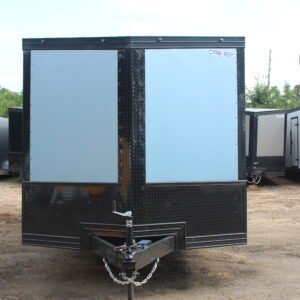 Front view of a light blue enclosed cargo trailer with a black diamond plate lower section and trim, featuring a "Cynergy" logo and A-frame tongue hitch, parked on a dirt lot surrounded by other trailers.