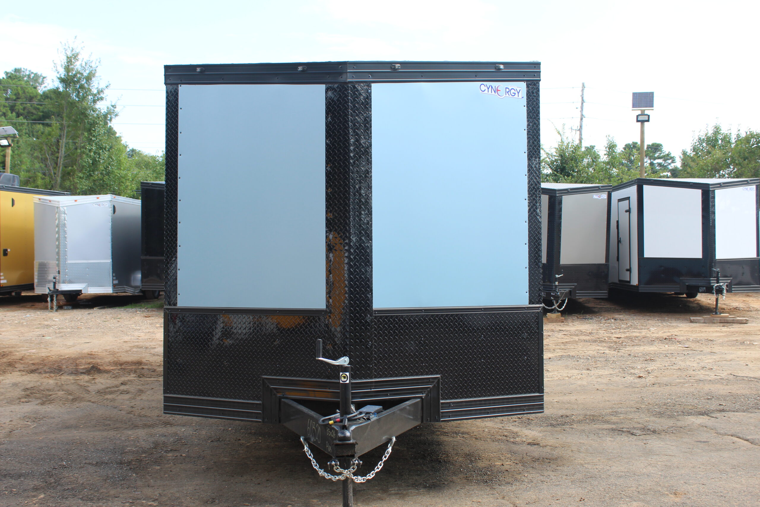 Front view of a light blue enclosed cargo trailer with a black diamond plate lower section and trim, featuring a "Cynergy" logo and A-frame tongue hitch, parked on a dirt lot surrounded by other trailers.