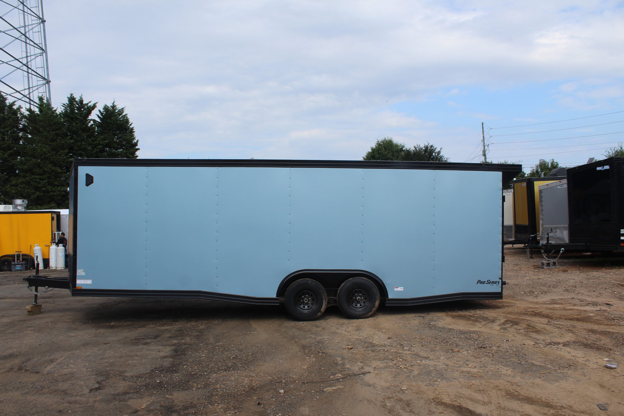 Side view of a light blue enclosed cargo trailer with black trim and tandem wheels, branded "Pro Series," parked on a dirt lot among other trailers under a cloudy sky.