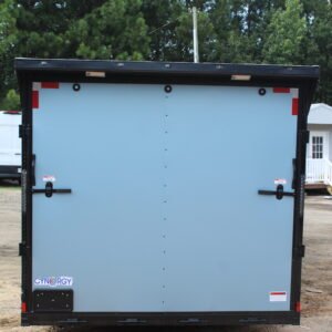 Rear view of a light blue enclosed cargo trailer with double barn doors, black trim, and red reflective safety markers. The trailer is branded "Cynergy" and parked on a dirt surface with trees and a small white building in the background.