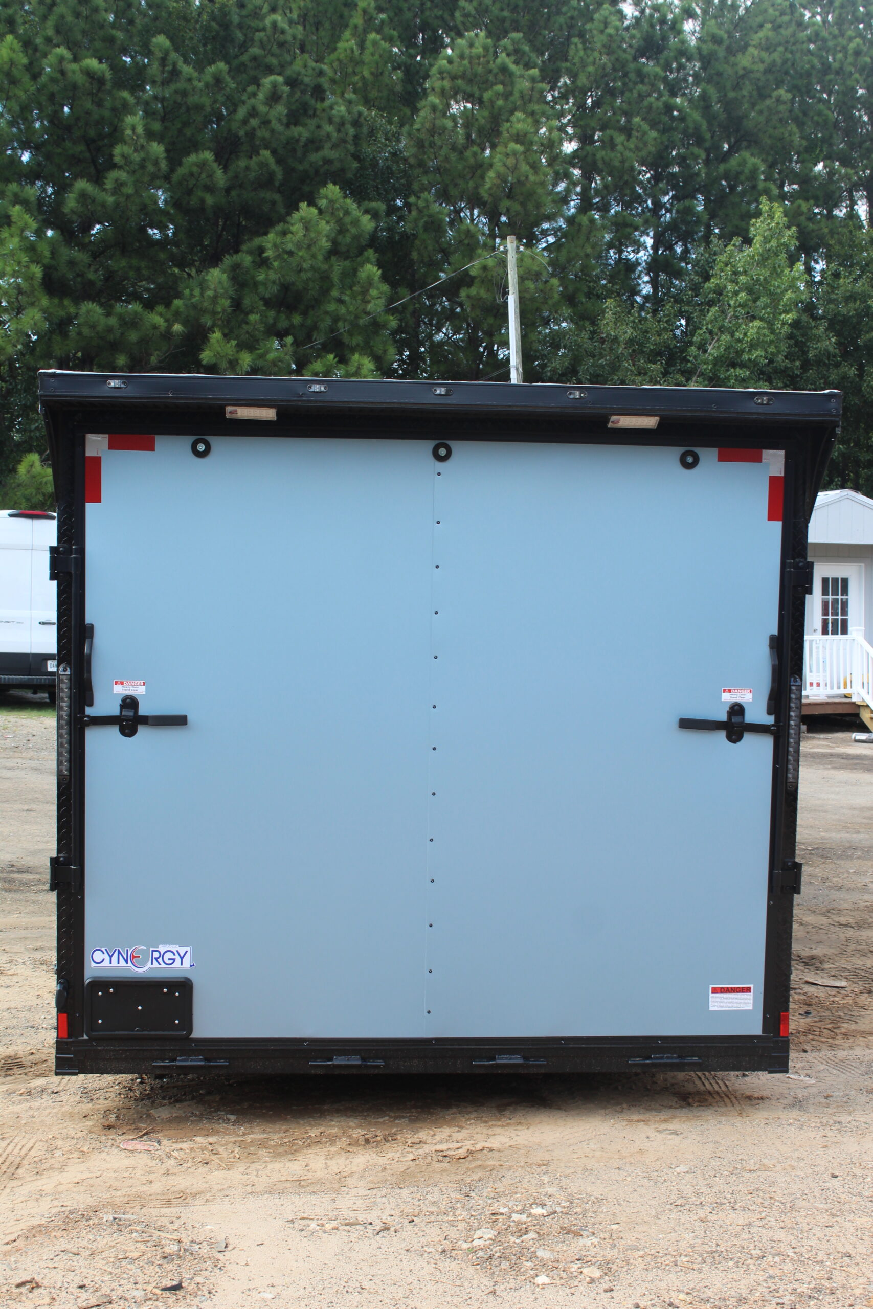 Rear view of a light blue enclosed cargo trailer with double barn doors, black trim, and red reflective safety markers. The trailer is branded "Cynergy" and parked on a dirt surface with trees and a small white building in the background.