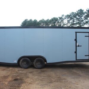 Side view of a large enclosed cargo trailer with a light blue exterior and black trim, featuring a single side door and tandem wheels, parked on a dirt lot with trees in the background.
