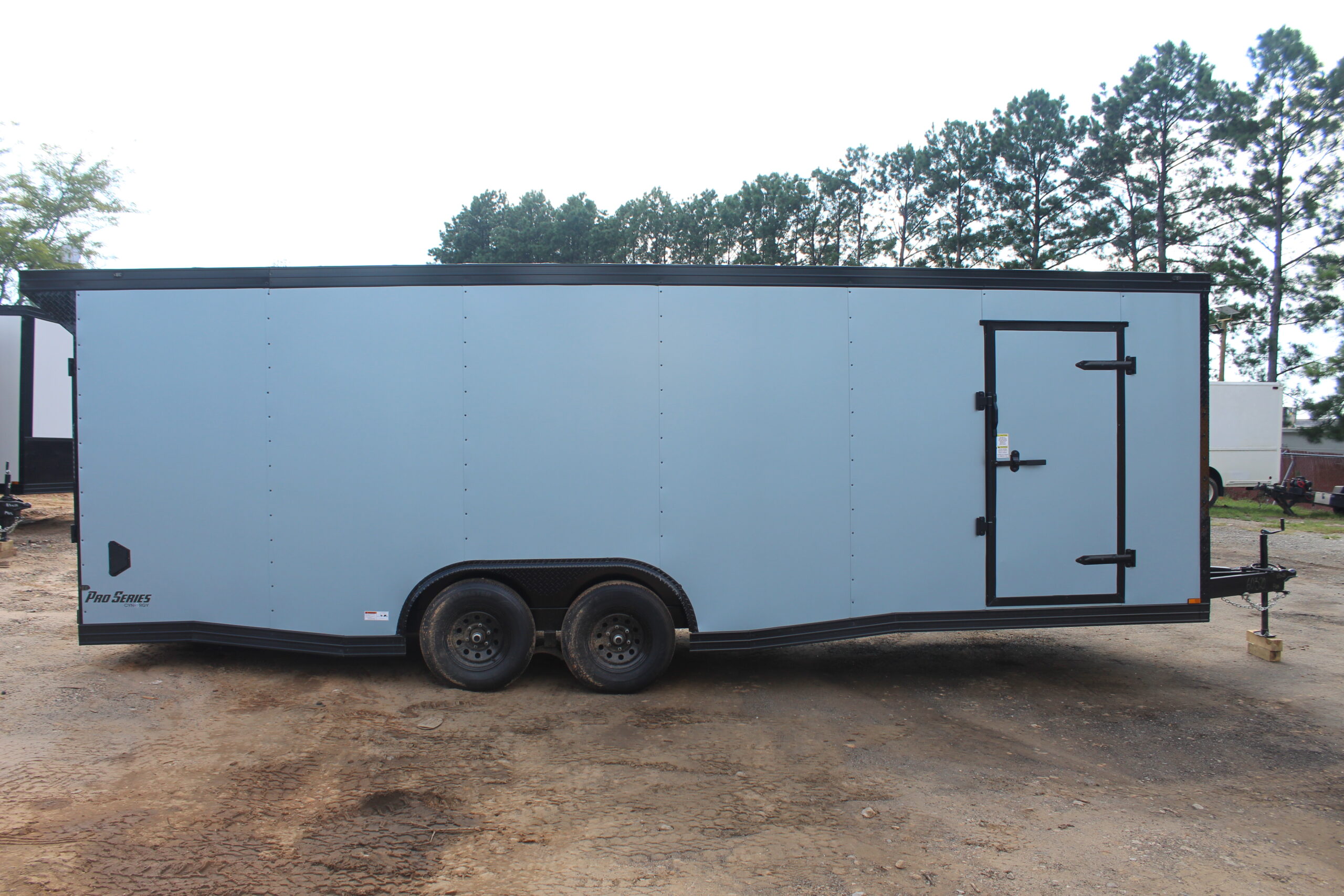 Side view of a large enclosed cargo trailer with a light blue exterior and black trim, featuring a single side door and tandem wheels, parked on a dirt lot with trees in the background.
